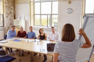 Frau steht neben Flipchart und, Kollegen am Meetingtisch schauen auf Flipchart